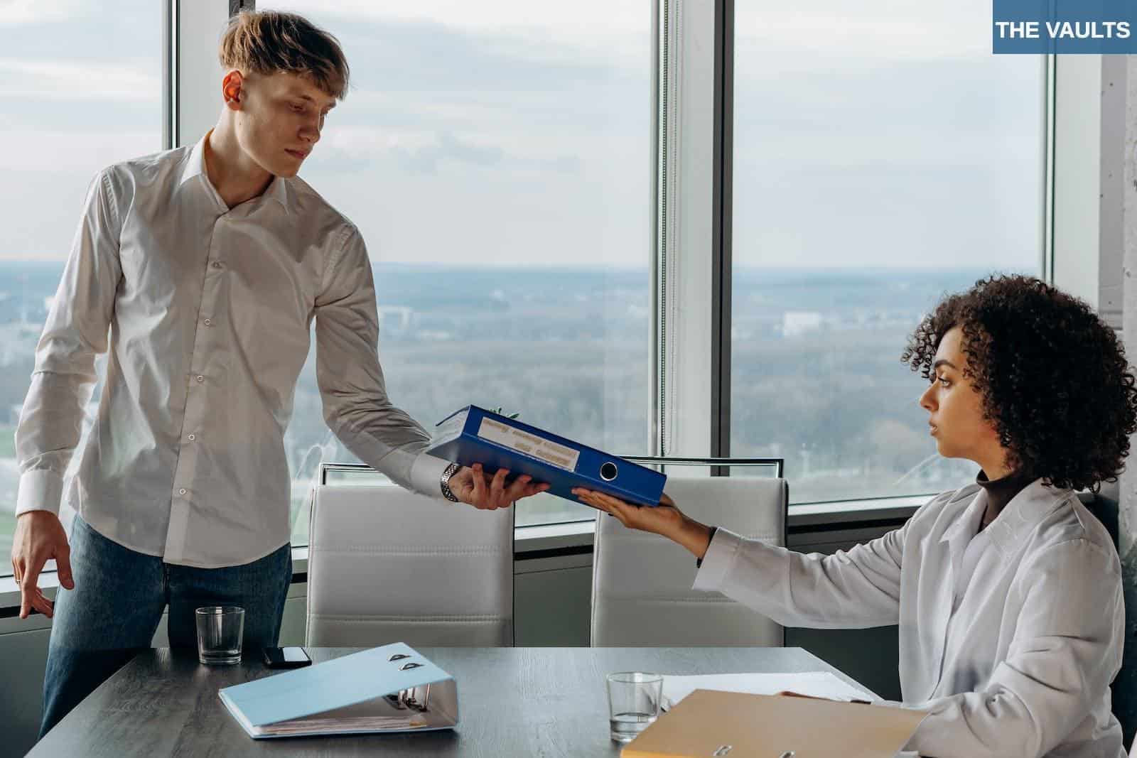 Young Man Standing in White Long Sleeve Shirt Giving a Book Report to His Coworker Inside a Conference Room