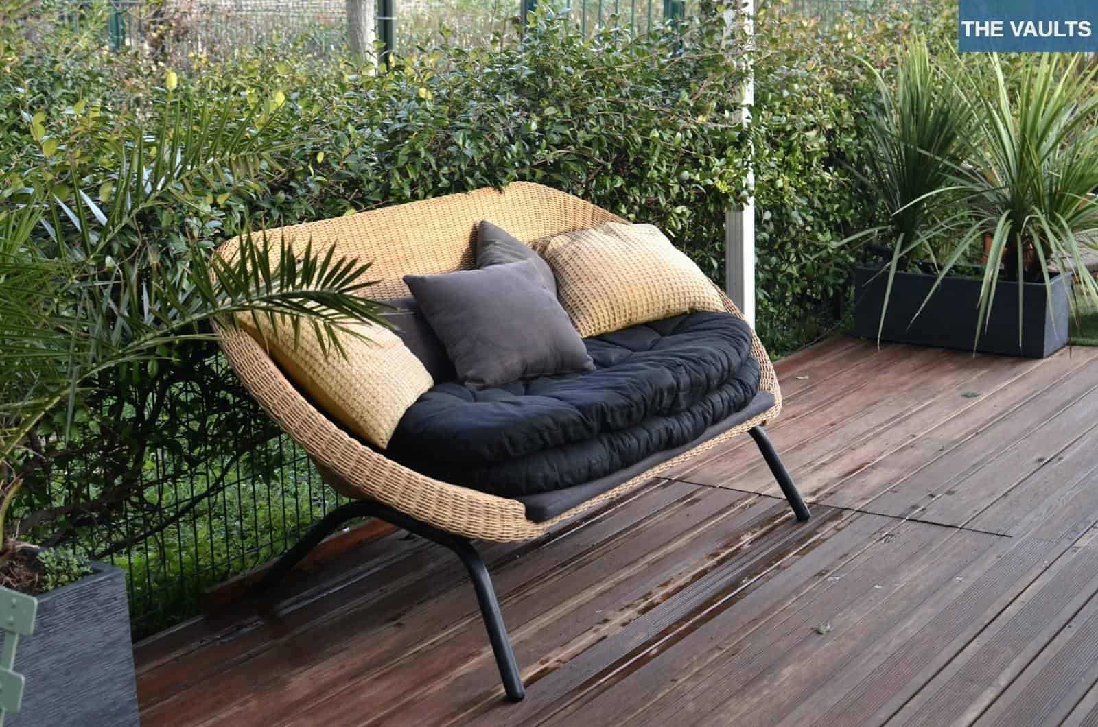 Comfortable stylish wicker sofa with soft seat and pillows placed in patio with lush green tropical vegetation in daytime